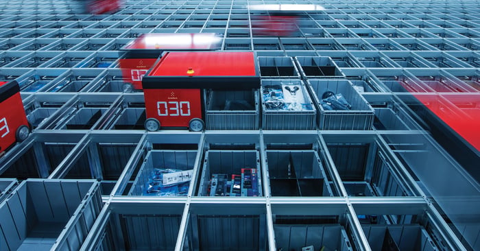 Red robots digging gray storage bins out of metal grid system in a warehouse  